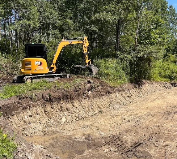 A tracked excavator digging a deep foundation trench on a residential lot in Lansing, Michigan, with exposed clay soil walls and a cleared site ready for footing work.